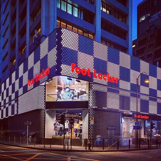 Exterior view of a Foot Locker store with a checkered pattern design at night in a city.