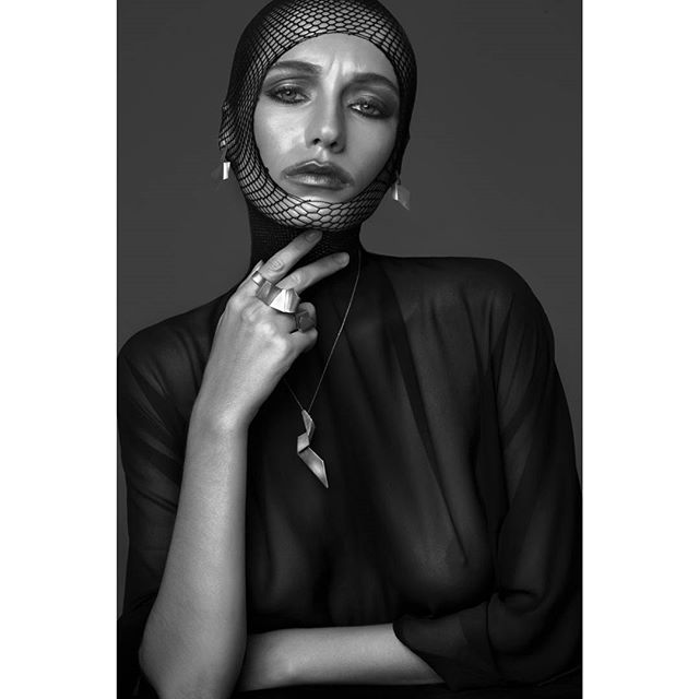 Monochrome portrait of a woman with a net head covering, silver jewelry, and a sheer top, exuding a mysterious and intense mood.
