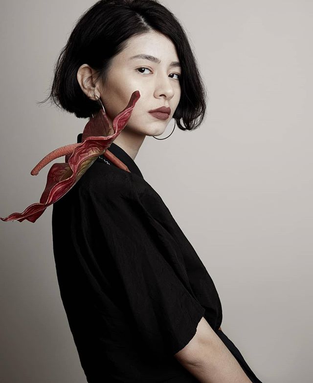 Portrait of a stylish woman with short dark hair, wearing an anthurium flower like a shawl and a black dress in a studio.