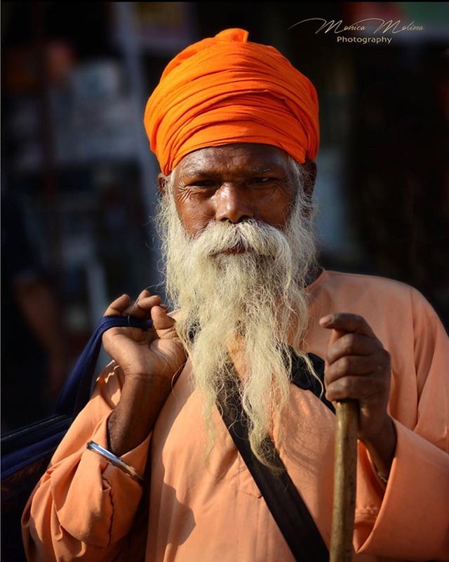 Portrait of an elderly Sikh man with a long white beard and orange turban, holding a cane.