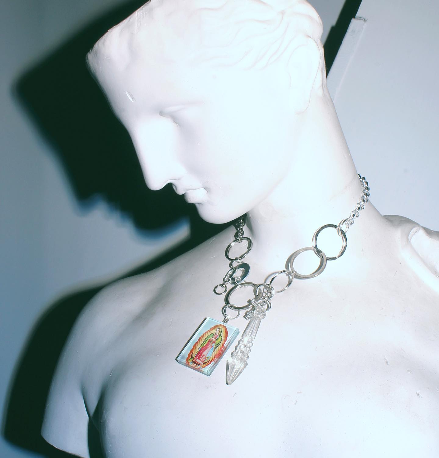 A bust statue is adorned with a silver chain necklace featuring a Virgin of Guadalupe pendant and crystal.