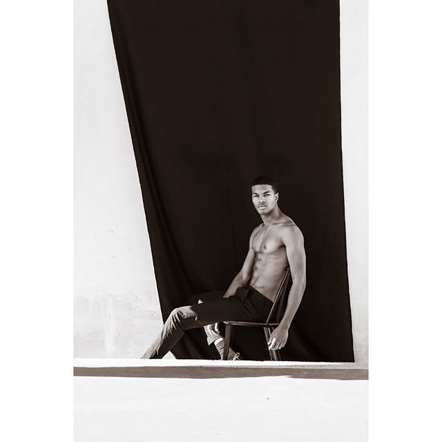 A shirtless man sits on a chair in a black and white studio shot, creating a moody and confident portrait.