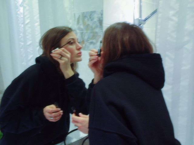 A woman in a black hoodie applies mascara in front of a bathroom mirror, getting ready for the day.