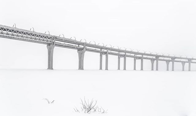 A long bridge stretches across a snow-covered landscape on a foggy winter day. 