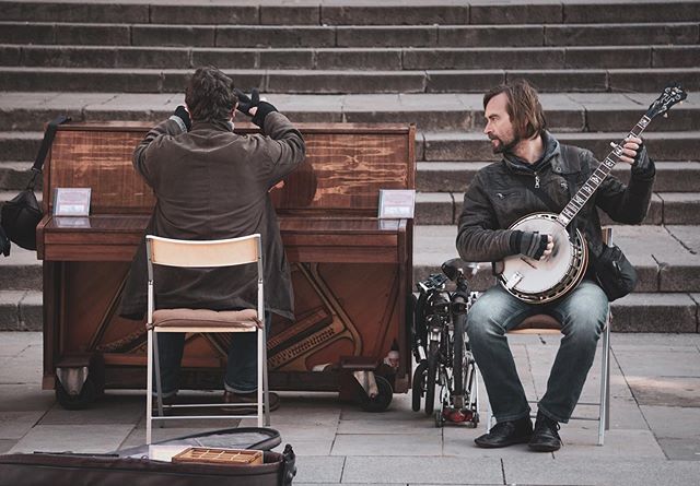 Two musicians play piano and banjo on city steps for passersby in an urban setting.