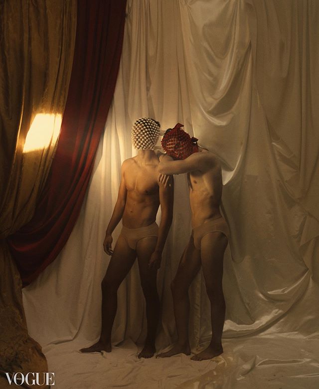 Two masked male figures pose artistically in front of a white backdrop for a fashion editorial.