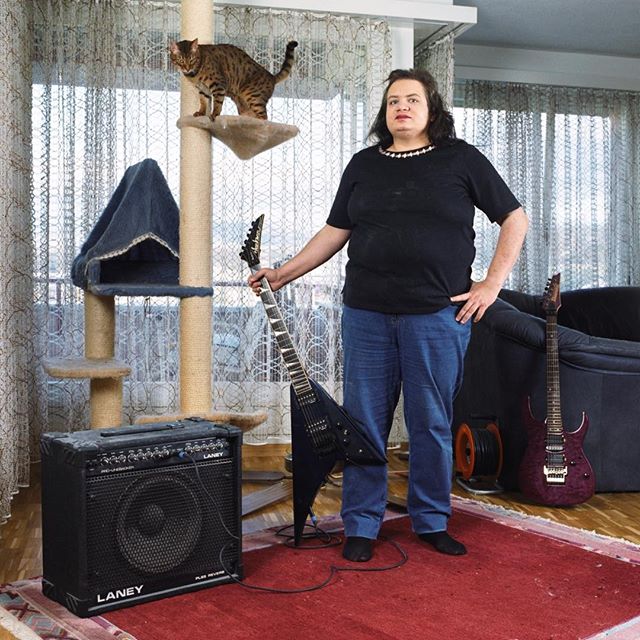 A man with a guitar poses in his living room alongside his cat and music equipment.