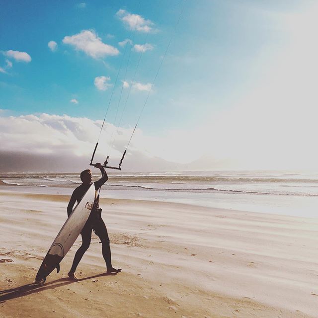 A kiteboarder carrying his board on a sandy beach, ready to ride in the ocean.