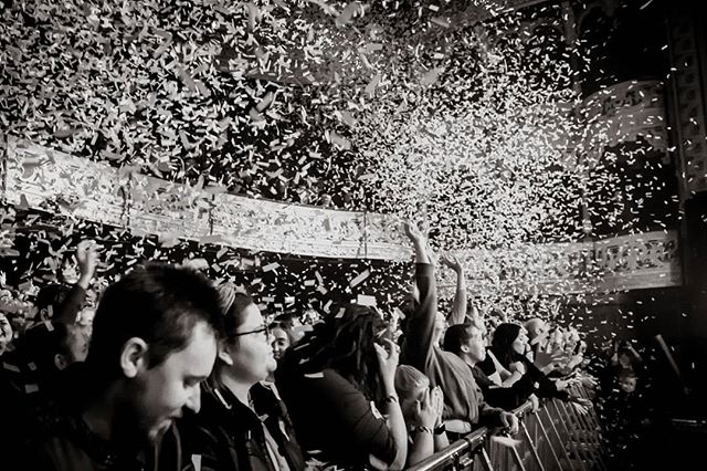 A black and white image captures the excitement of a live concert with a crowd under a shower of confetti.