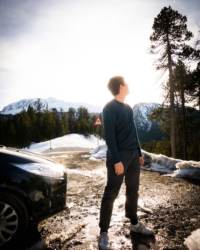 A man stands near his car on a snowy road, looking up at the mountains on a bright day.
