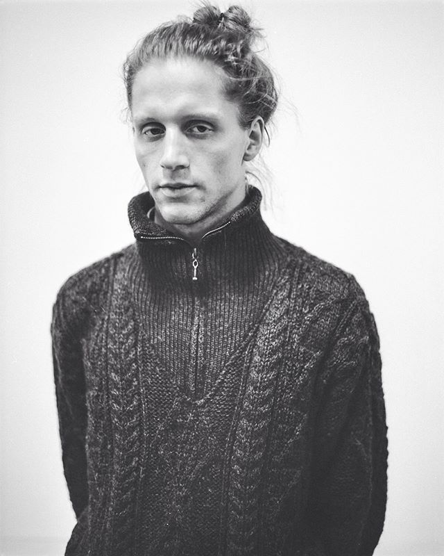 A black and white portrait of a man in a knit sweater against a neutral backdrop.