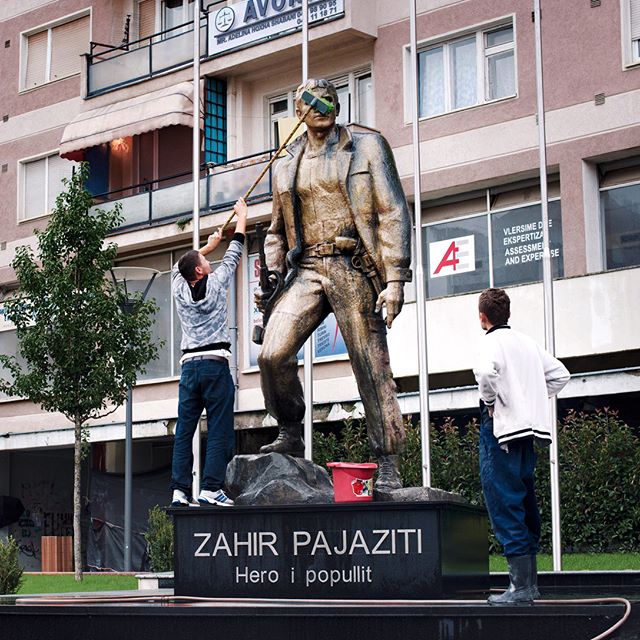 Two men cleaning a statue of Zahir Pajaziti in a city setting, focusing on maintenance and public art.