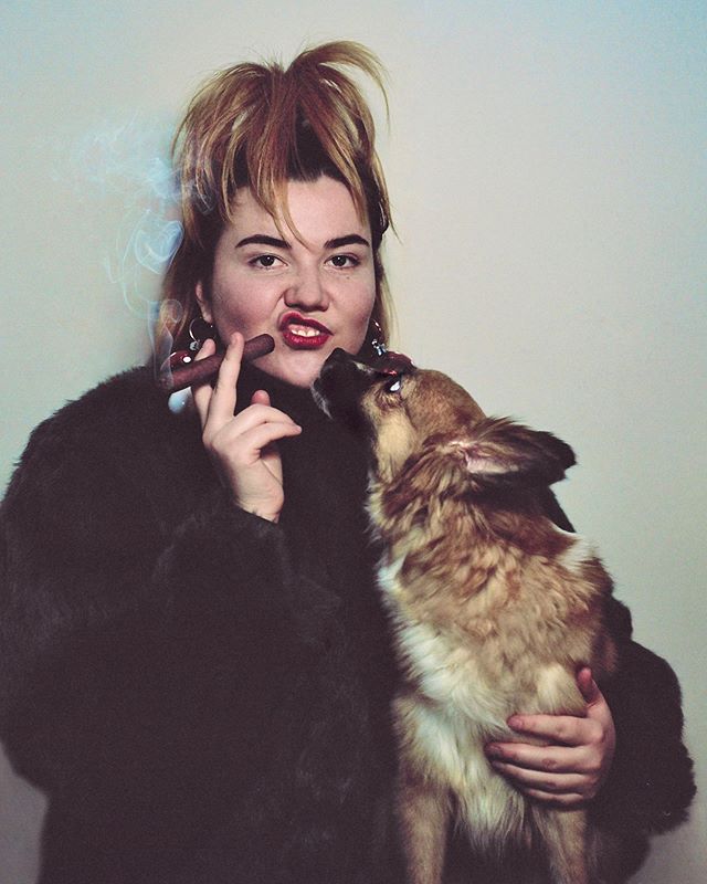 A woman in a fur coat holds a dog while smoking a cigar. A playful, editorial-style shot for social media.