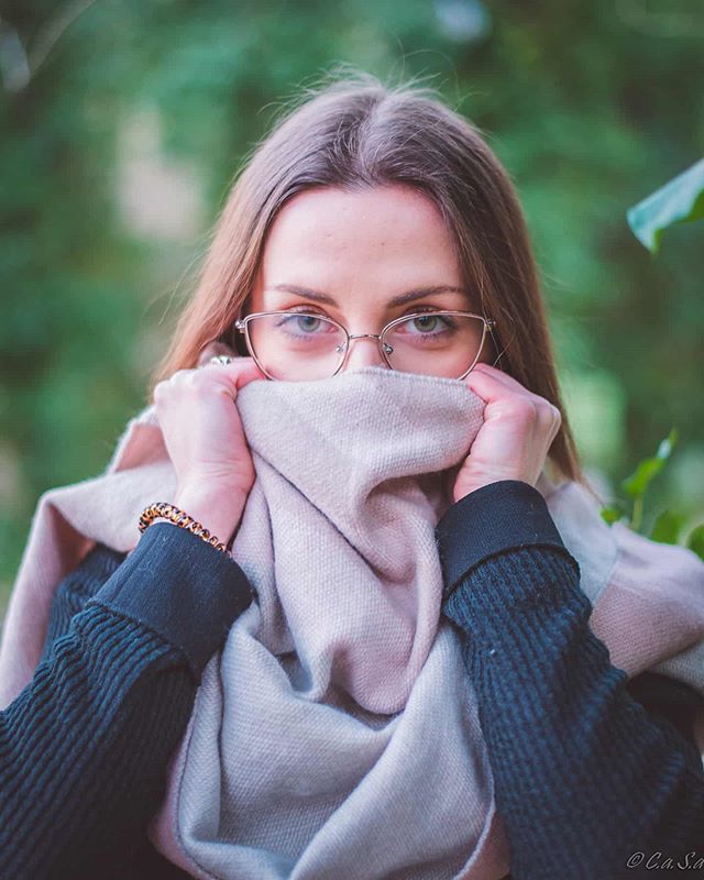 A young woman with glasses covers her face with a scarf, creating a cozy and mysterious portrait in a natural outdoor setting.
