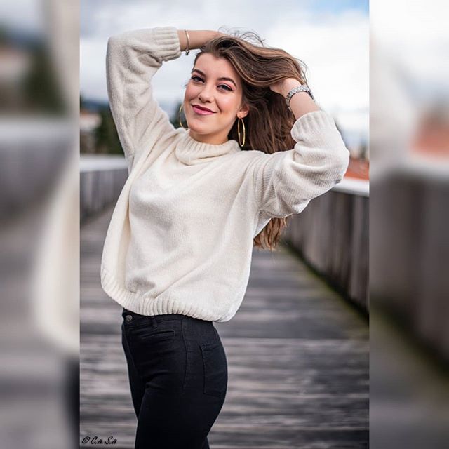 A smiling brunette woman in a sweater poses outdoors. She has hoop earrings and dark pants.