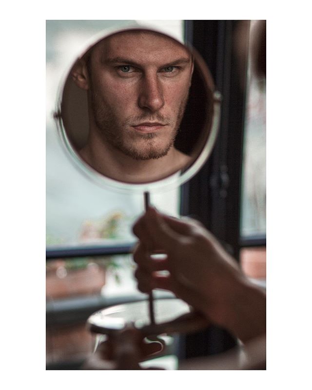 A serious man gazes from a round mirror in a soft, naturally lit indoor setting.