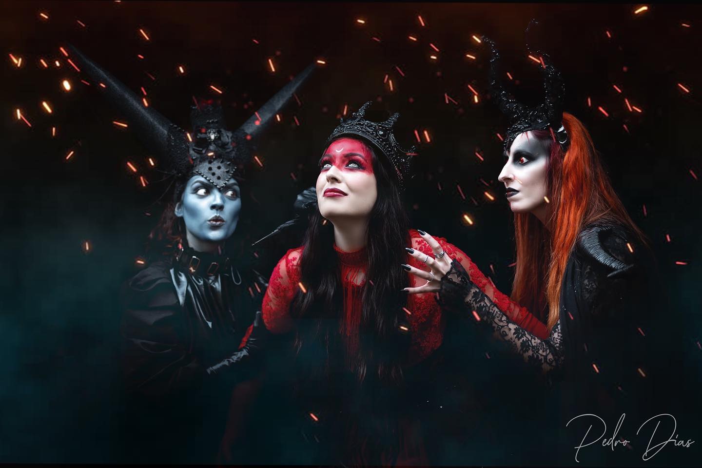Three women in fantasy costumes pose in a dark setting with soft lighting and intense makeup.