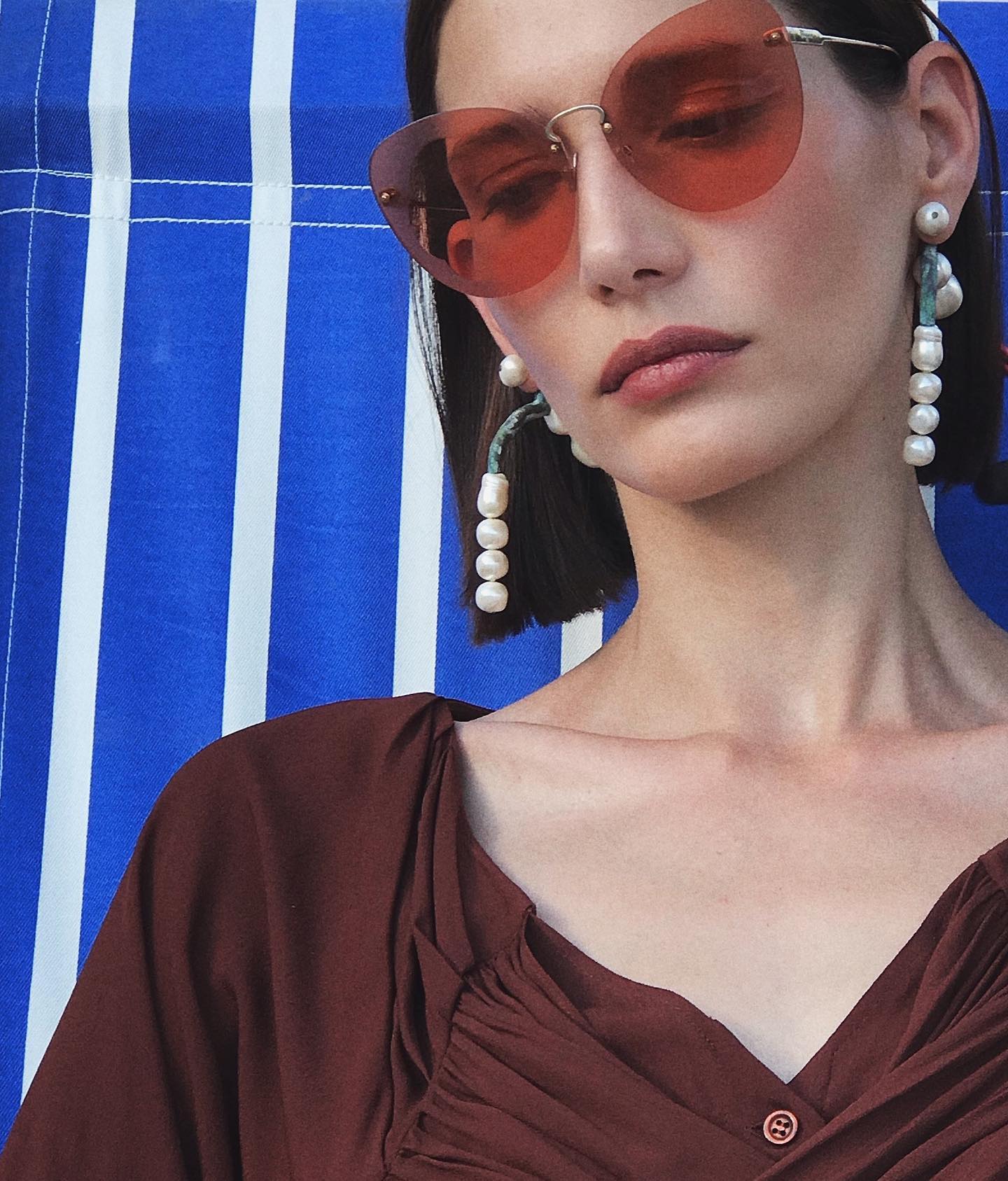 A woman wearing sunglasses and pearl earrings poses against a blue and white striped background.