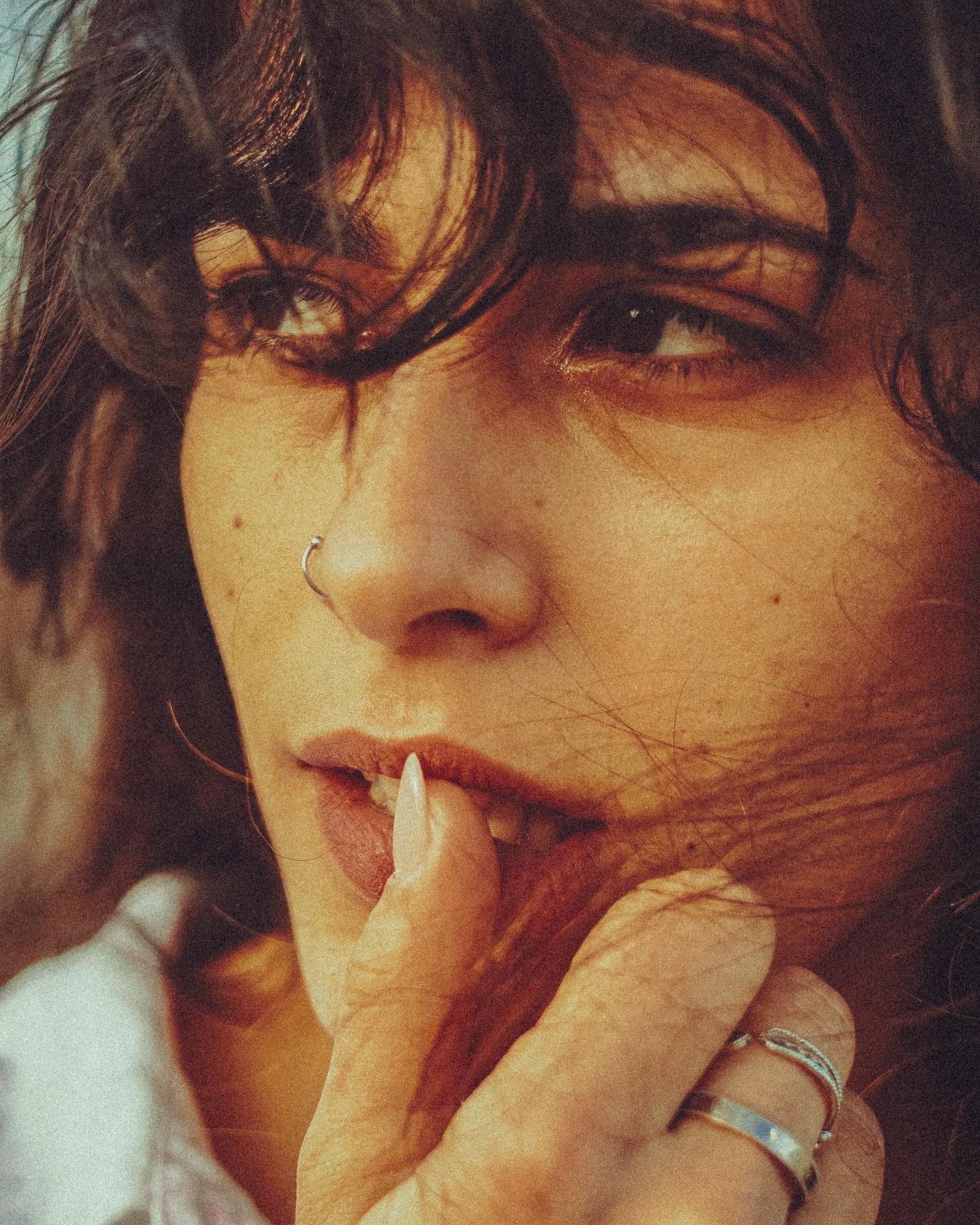 Close-up of a woman with a nose ring and multiple finger rings posing with fingers near her mouth.