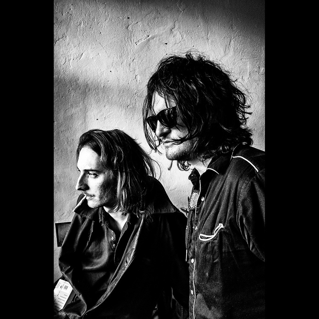 A moody black and white portrait of two men with long hair, one smoking, in a vintage editorial style.