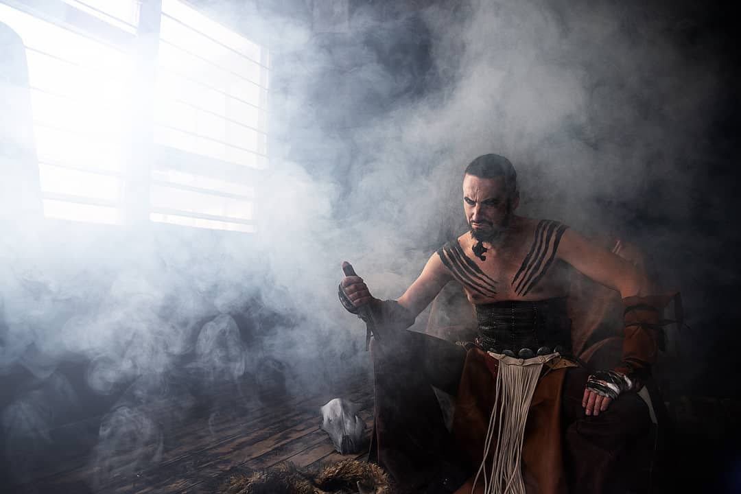 A man in warrior costume poses in a smoke-filled room, creating a dramatic and powerful image.
