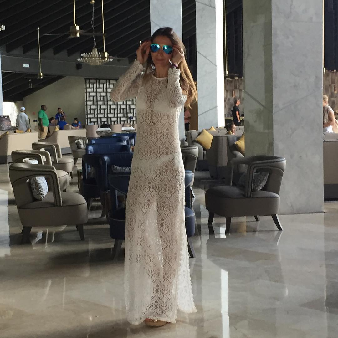 A woman in a long white lace dress and sunglasses stands in a hotel lobby.