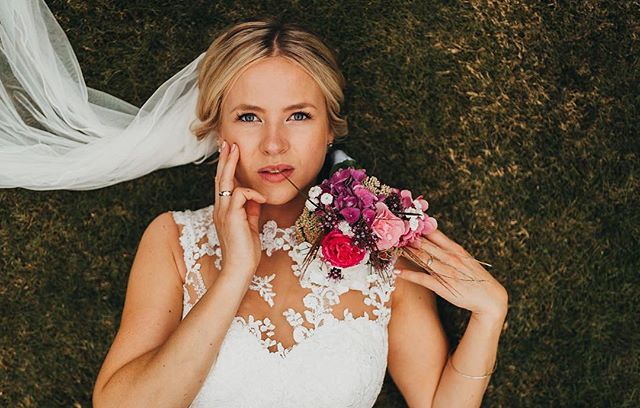 A bride lies on the grass in her wedding dress, holding a bouquet, in a dreamy outdoor setting.