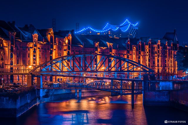 A night scene of illuminated buildings and a bridge reflected in the water of a city canal.