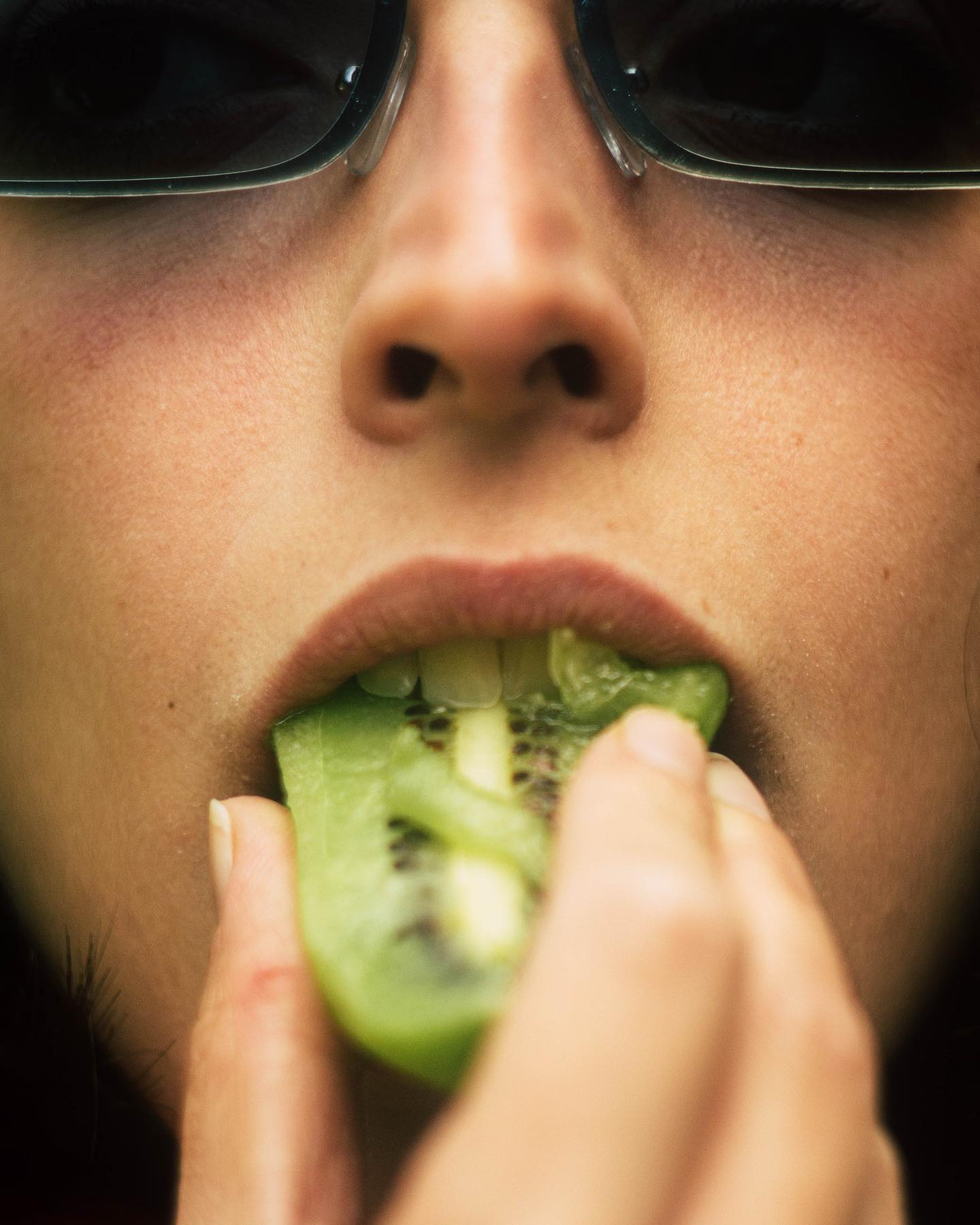 Close up of a woman eating a green kiwi slice, wearing sunglasses.
