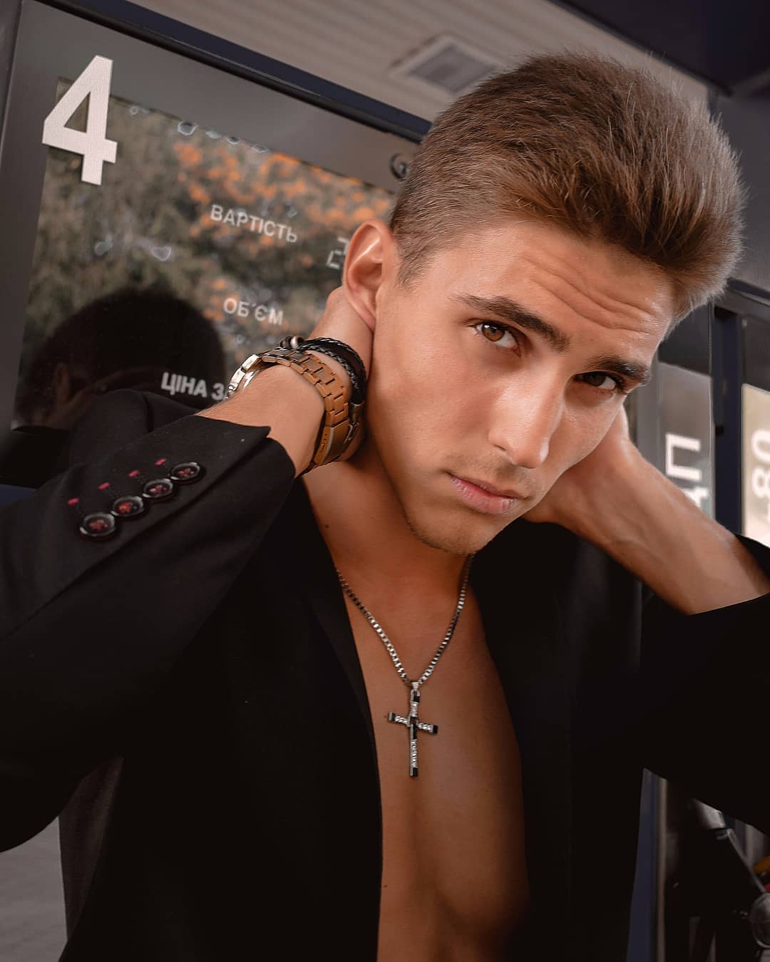 A handsome man poses in a black suit jacket with a cross necklace at a gas station, creating a stylish and confident image.