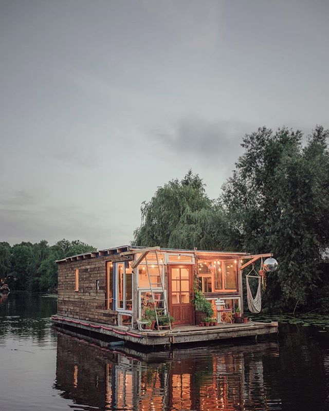 A charming wooden houseboat with plants and a hammock, reflecting on a calm lake surrounded by trees.