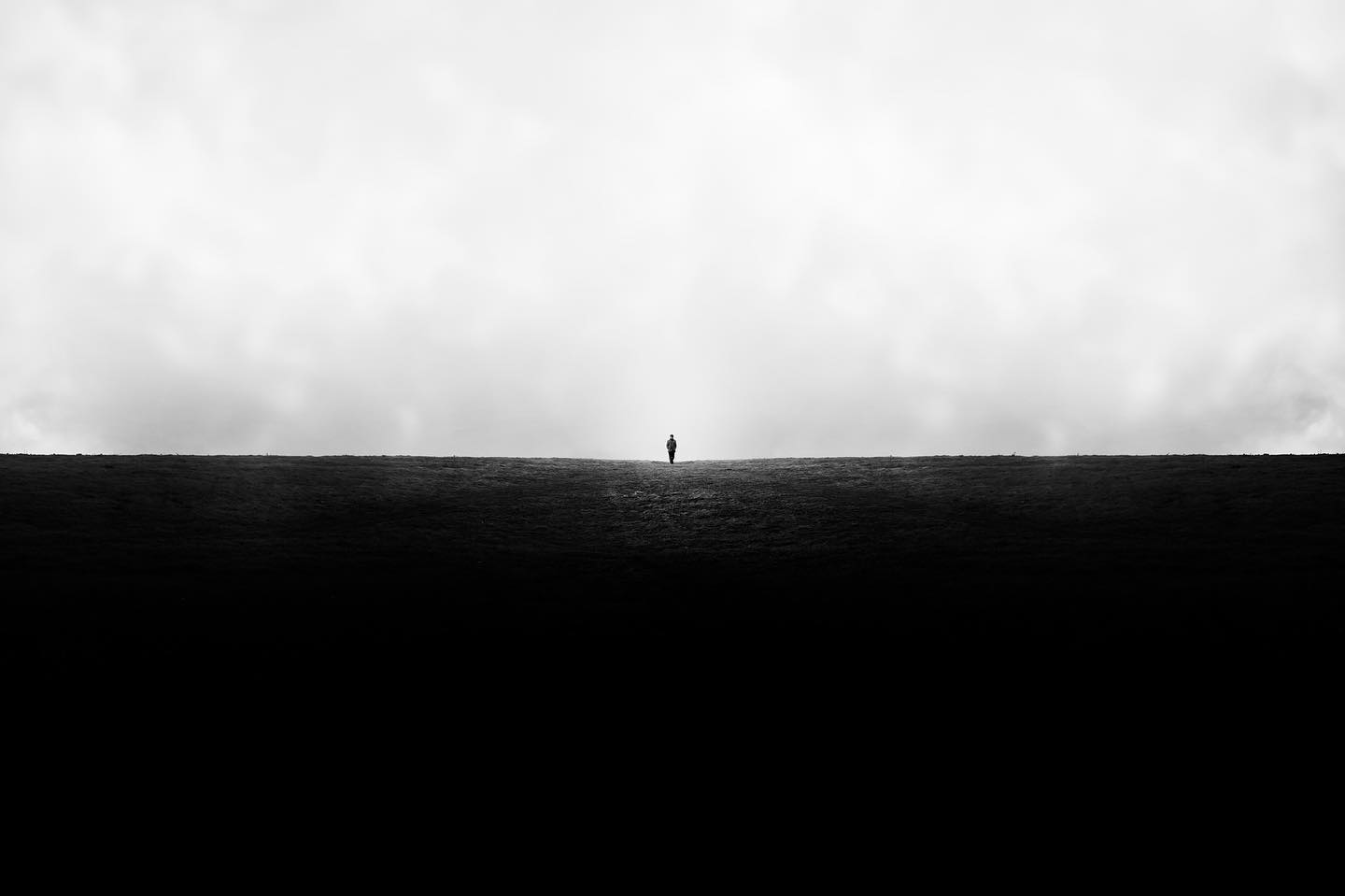 A lone figure walks on a dark field towards a cloudy horizon in a minimalist black and white landscape.