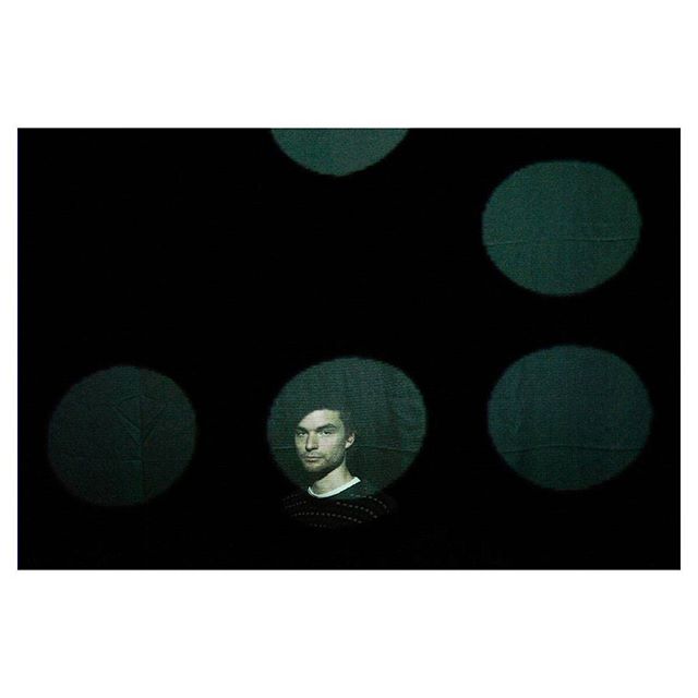 A man stands amidst a dark backdrop illuminated by soft, circular spotlights for a modern studio portrait.