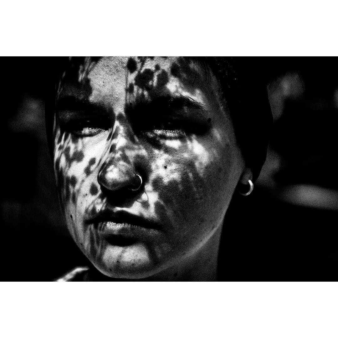 A black and white portrait of a woman's face with dappled light and shadows creating a mysterious and moody atmosphere.