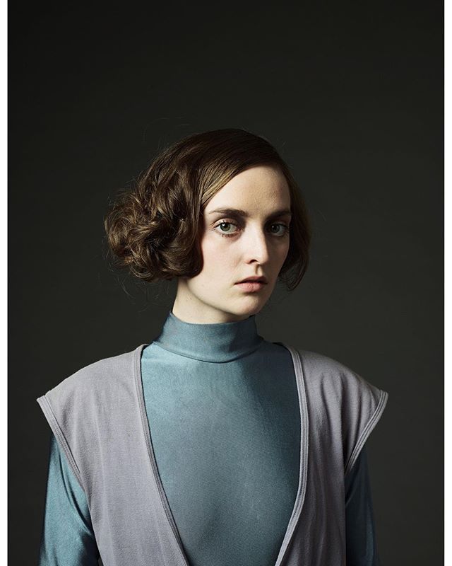 A brunette woman in a blue turtleneck and grey vest poses with a serious expression in a studio setting.
