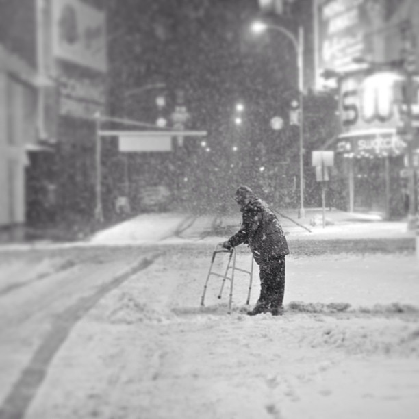 An elderly person uses a walker on a snowy city street at night in a black and white image.