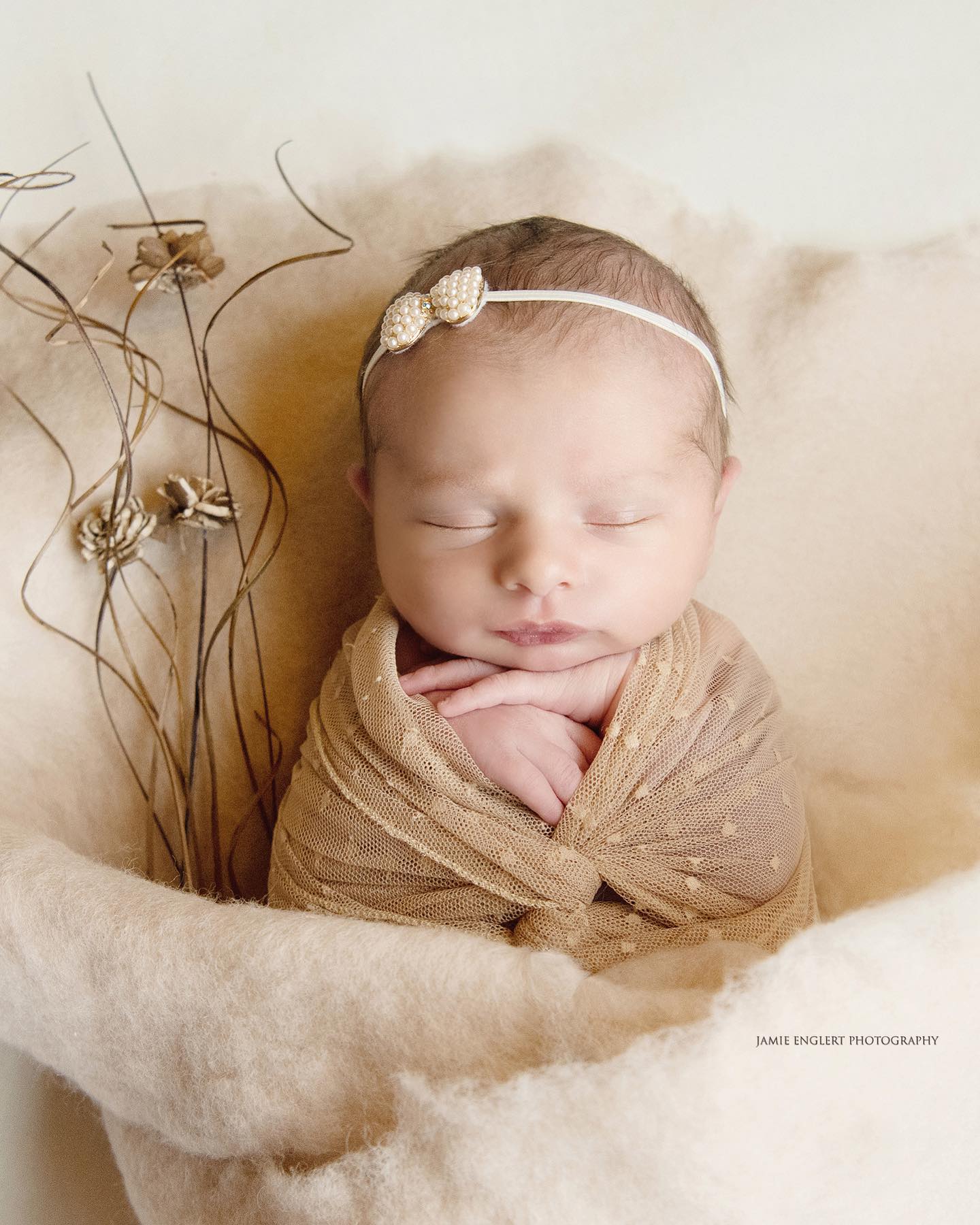 A sleeping newborn baby wrapped in a beige blanket with a pearl headband, photographed in a soft, warm setting.