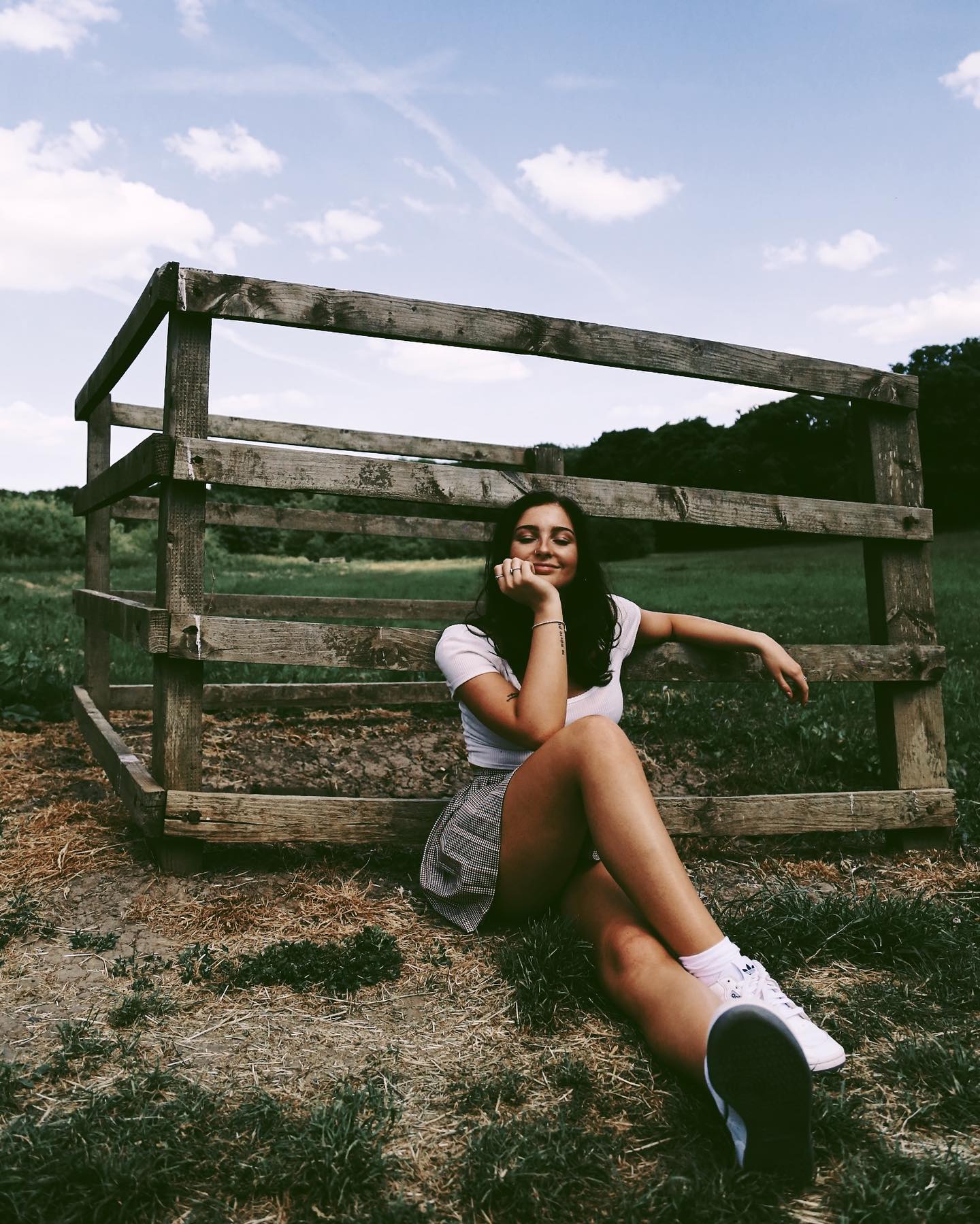 A brunette woman sits by a wooden fence in a field on a sunny day.