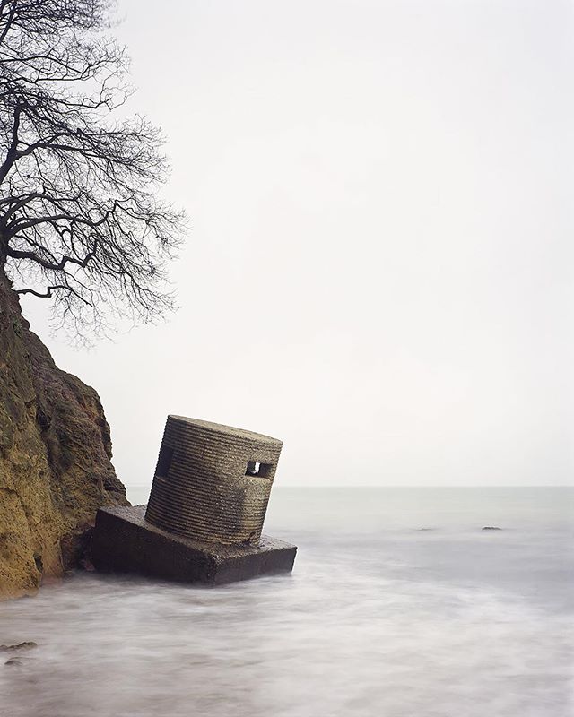 A historic pillbox sits on a beach, enduring coastal erosion under a cloudy sky in a tranquil scene.