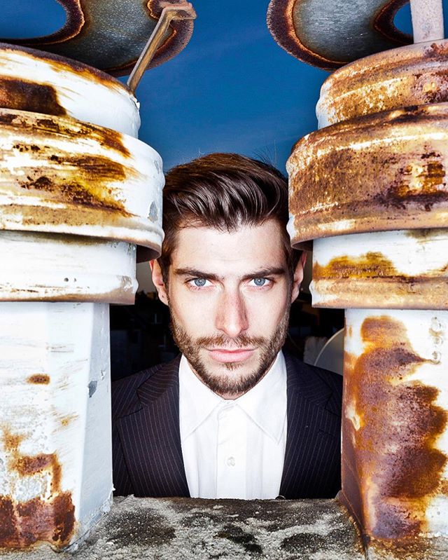 A handsome man with blue eyes poses confidently between weathered industrial objects in a striking portrait.