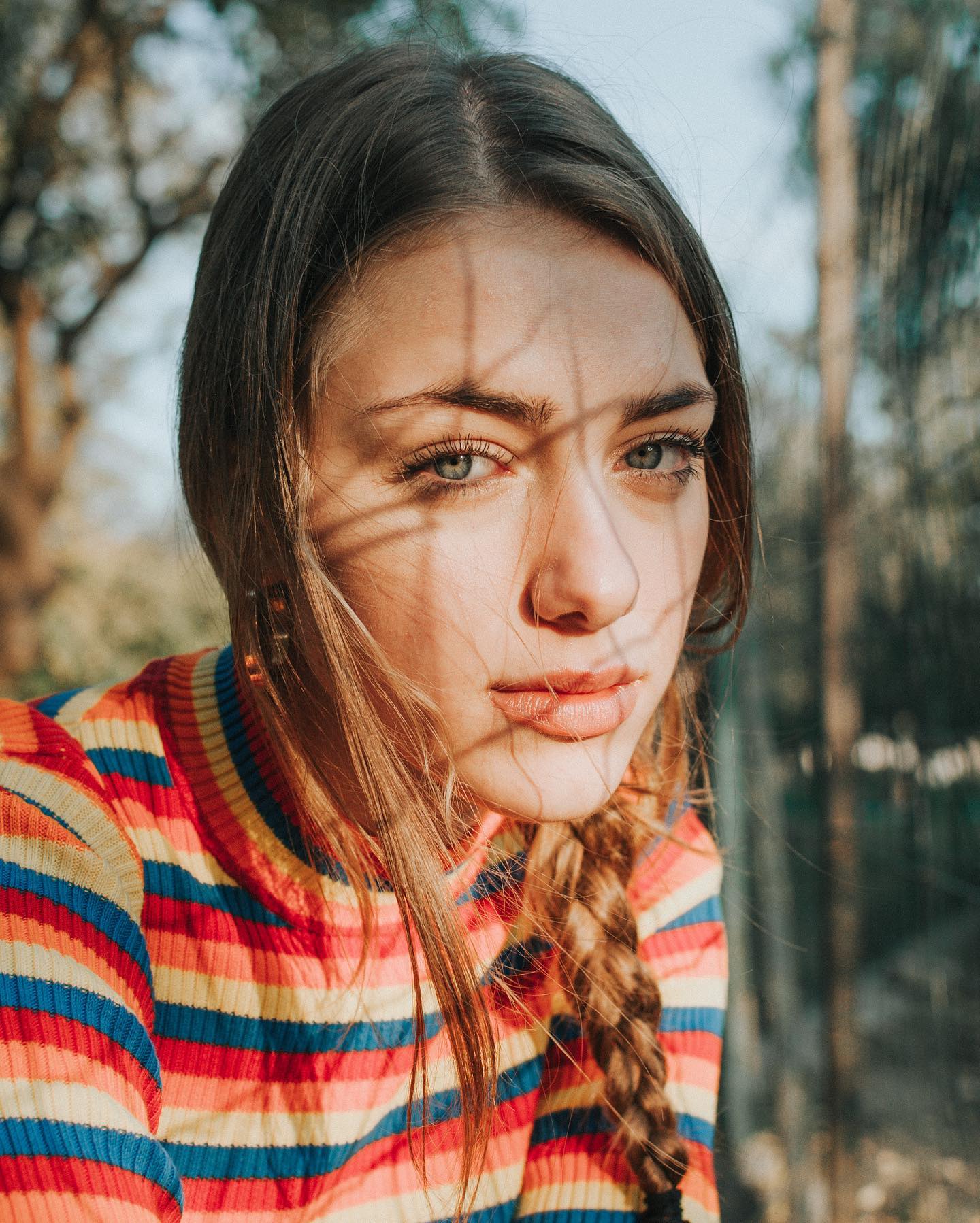 A young woman gazes dreamily into the camera, her face partially shadowed by branches in soft, natural light.