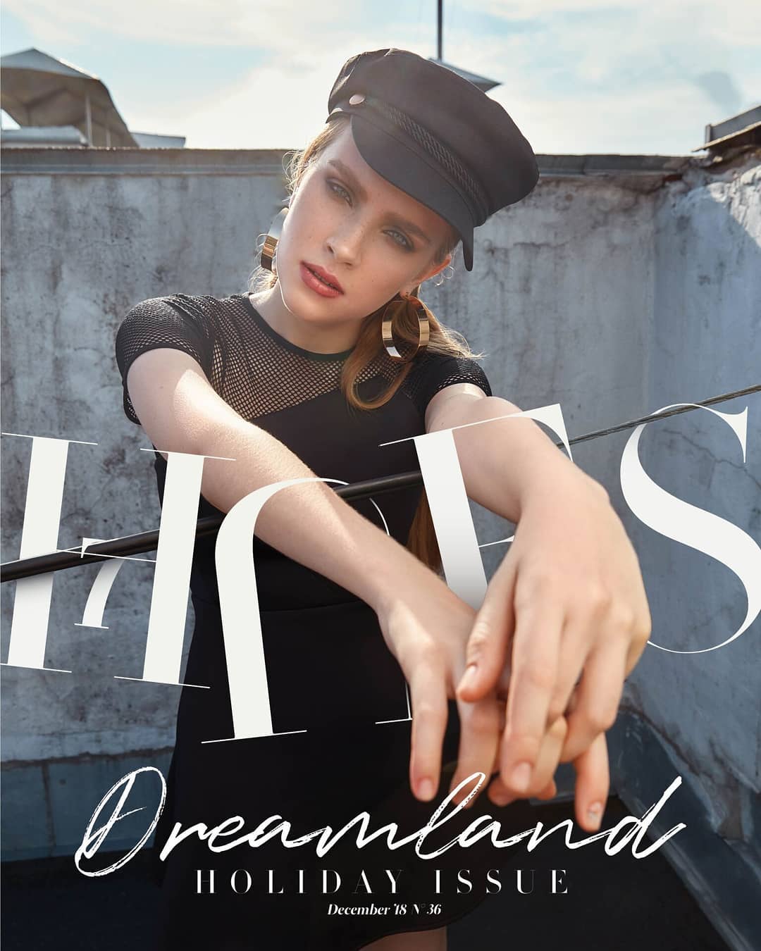 A stylish woman poses on a rooftop in a black dress, earrings, and hat for a fashion editorial.