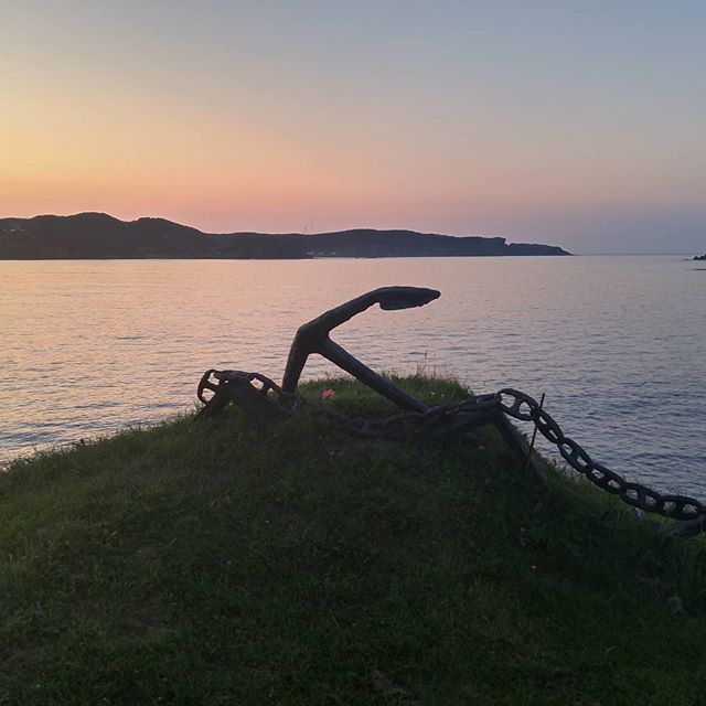An anchor and chain silhouette on a grassy hill overlook the calm ocean at sunset.