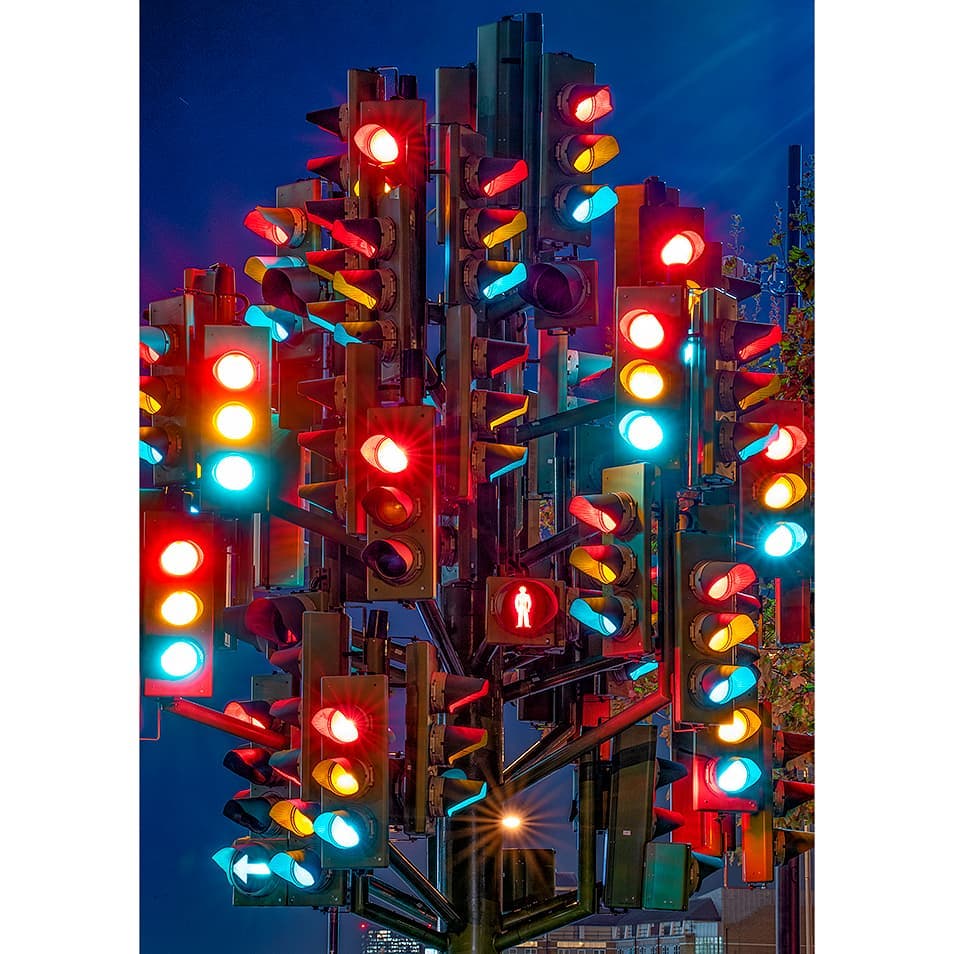 A whimsical sculpture of numerous traffic lights illuminates the city with vibrant colors against a deep blue evening sky.