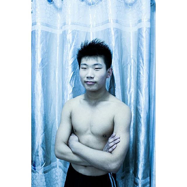 Shirtless man poses with crossed arms against a light blue curtain.