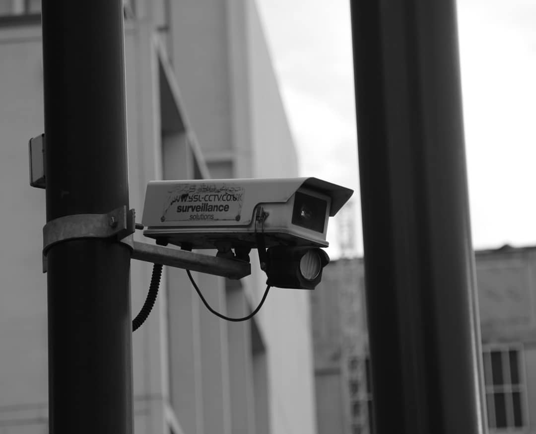A grayscale shot capturing a security camera mounted on a pole in an urban setting, emphasizing surveillance.