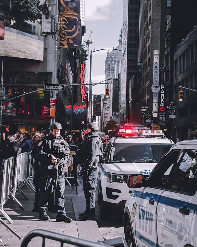 Two NYPD officers stand guard on a busy New York City street with patrol cars and towering buildings.