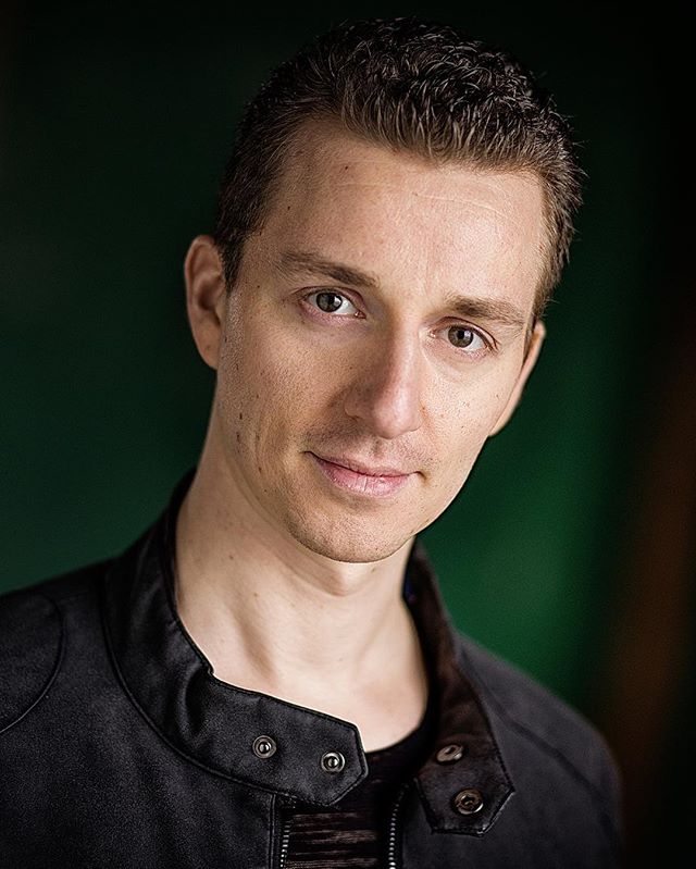 A portrait of a confident man wearing a black jacket against a green background.