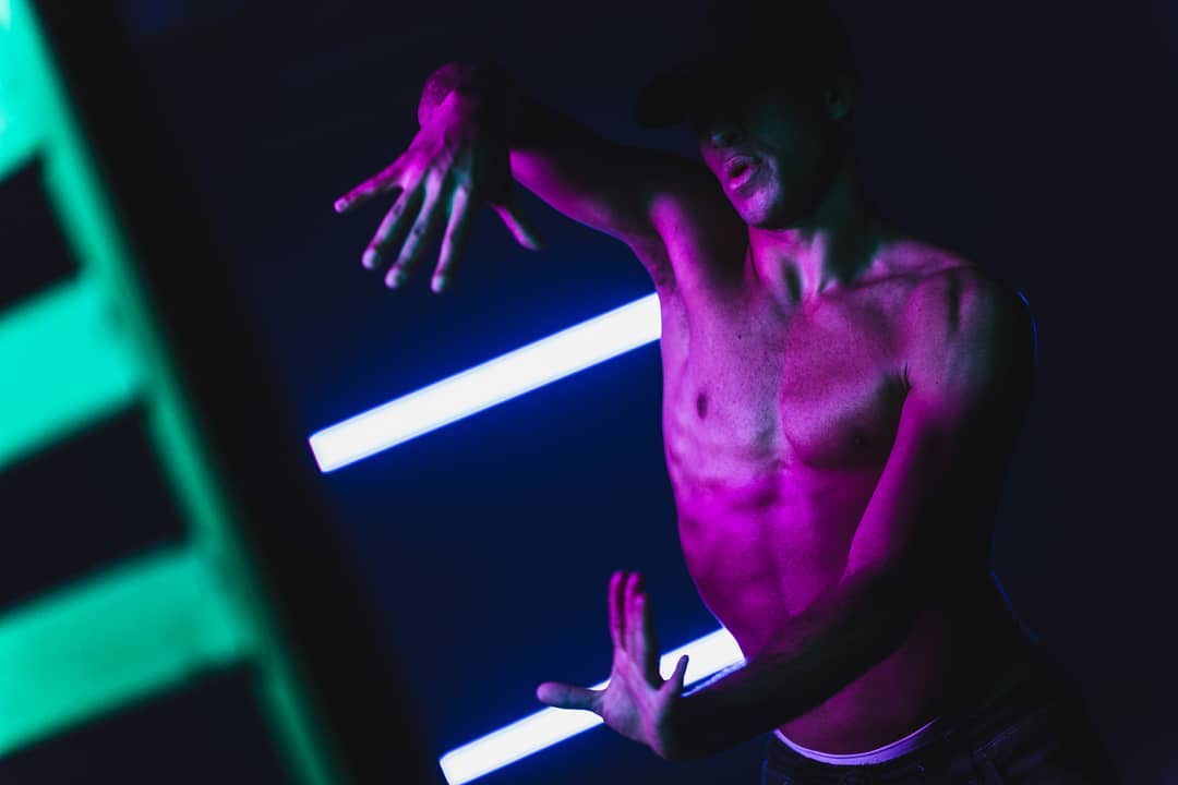 A shirtless male dancer performs under vibrant neon lighting in a dynamic studio shot.