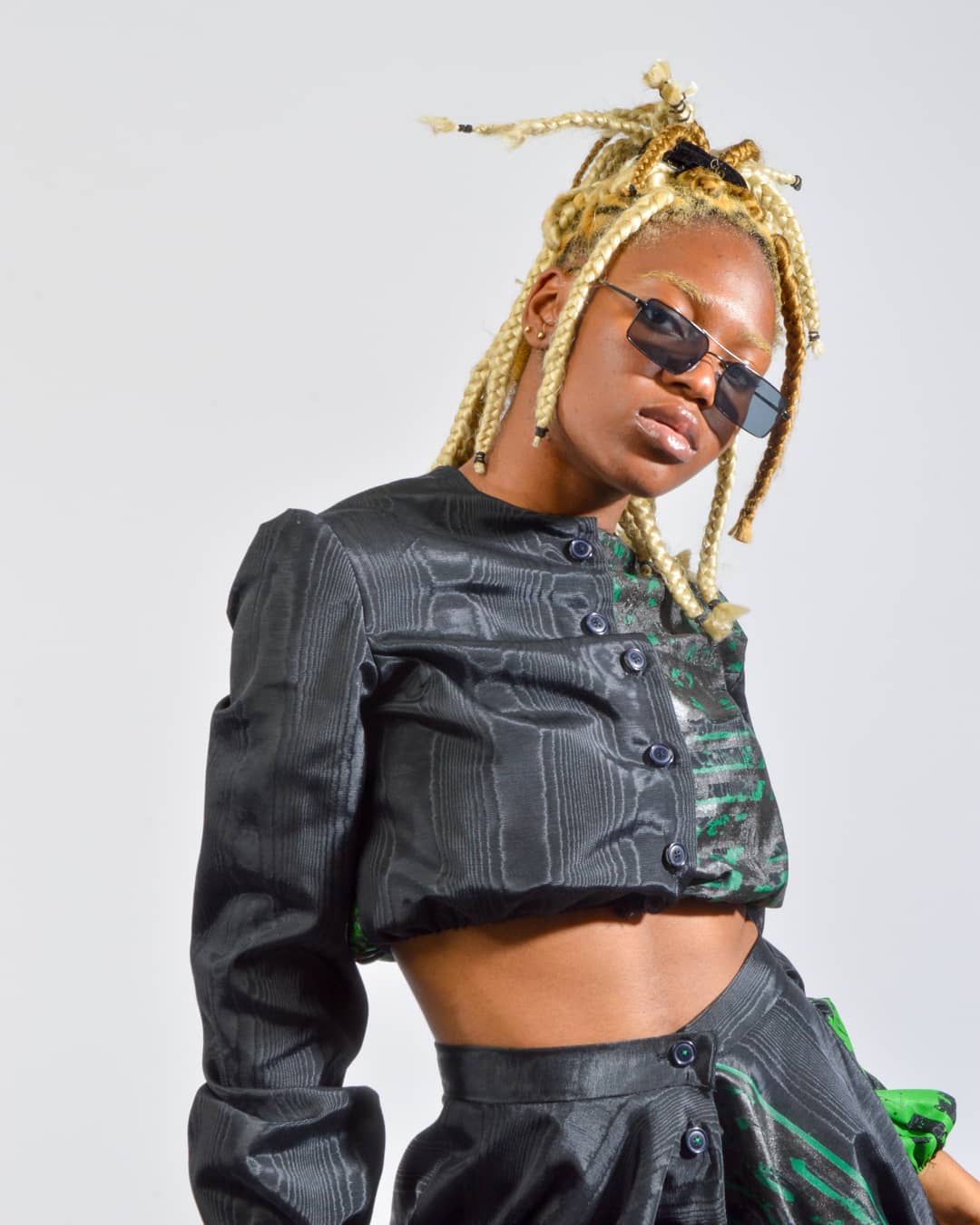 A stylish woman with blonde braids and sunglasses poses in a black and green patterned crop top and skirt.